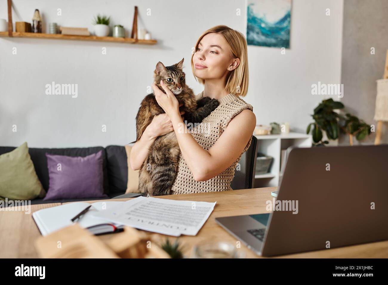 A serene woman with short hair sitting at a table, gently holding a cat in her arms at home. Stock Photo