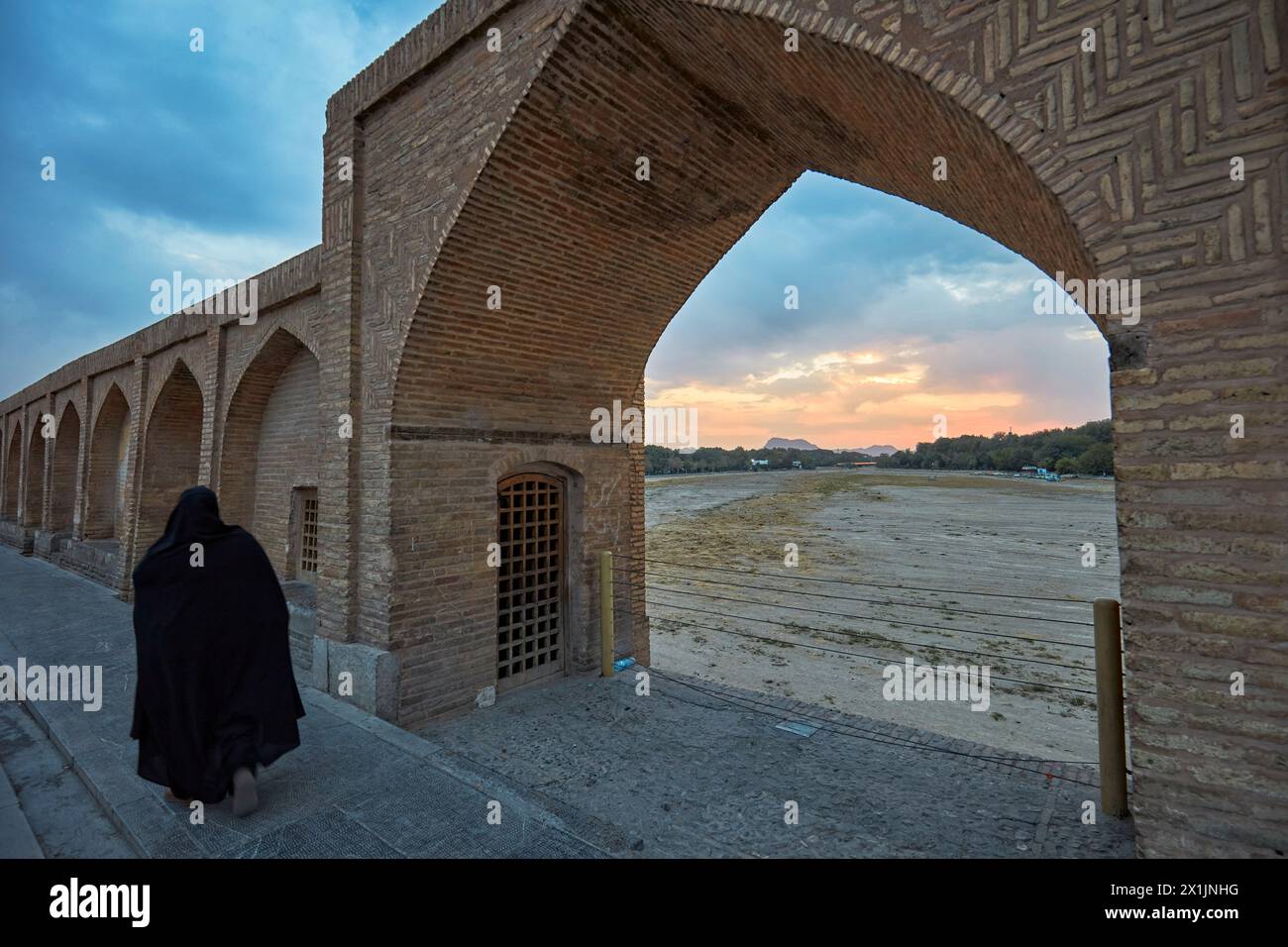 Iranian woman in black chador pass by an arch on the Si-o-se-pol Bridge with a view of dry Zayanderud river bed during dry season. Isfahan, Iran. Stock Photo