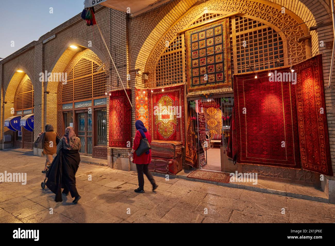 People walk by a handicraft shop in Naqsh-e Jahan Square with a selection of handmade Persian carpets displayed at the shopfront. Isfahan, Iran. Stock Photo