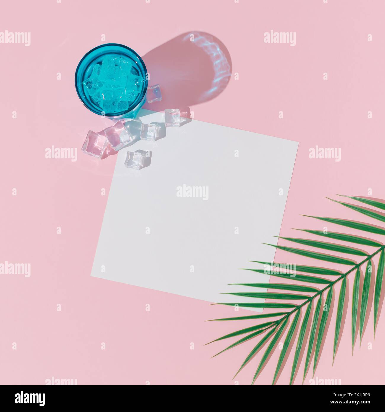 Creative summer layout made with cold refreshing drink, ice cubes, tropical palm leaf and paper card note copy space on pastel pink background. Stock Photo