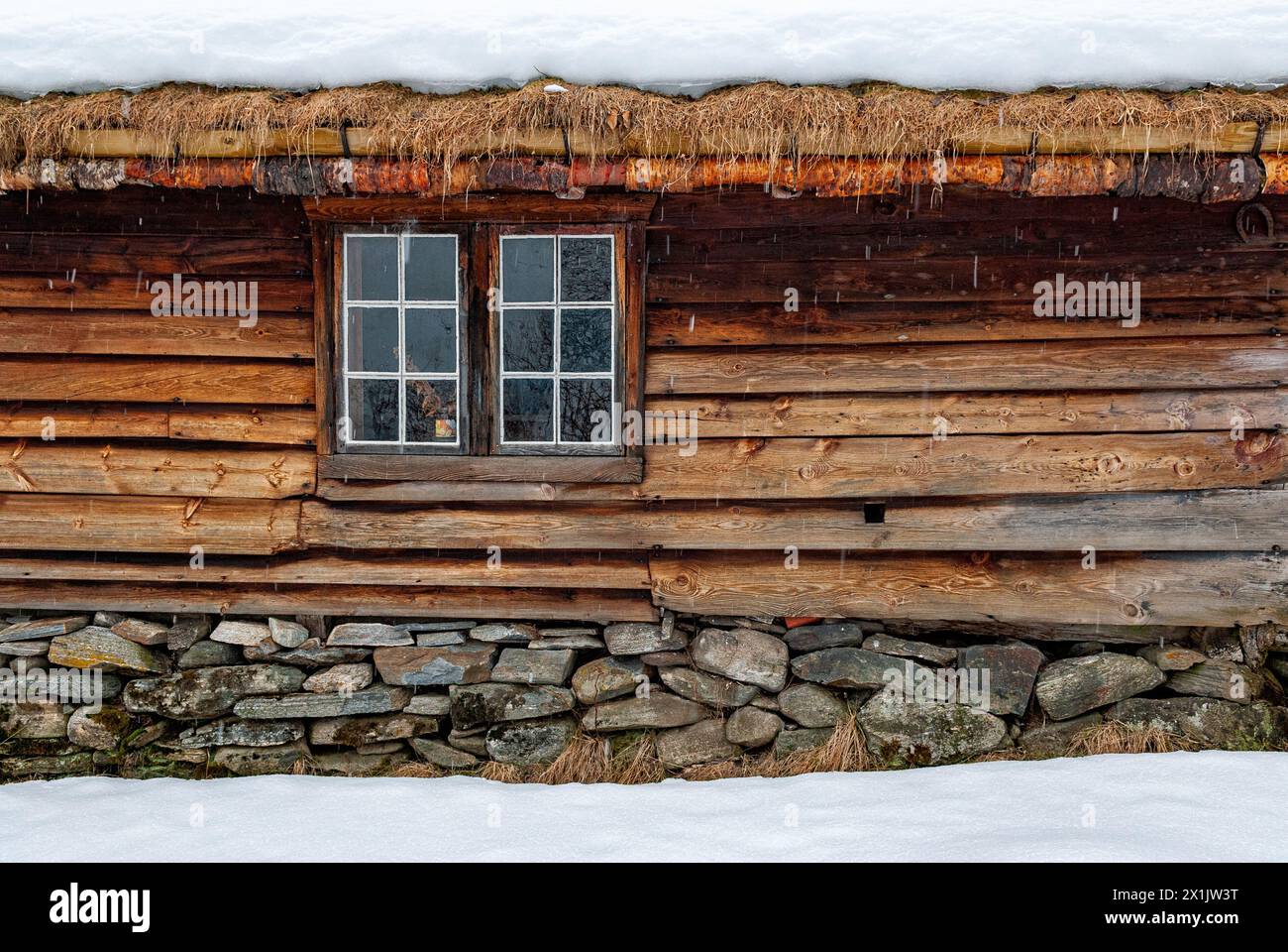 A rustic log cabins wall with a single window, thatched roof covered in snow, and stone foundation. Stock Photo