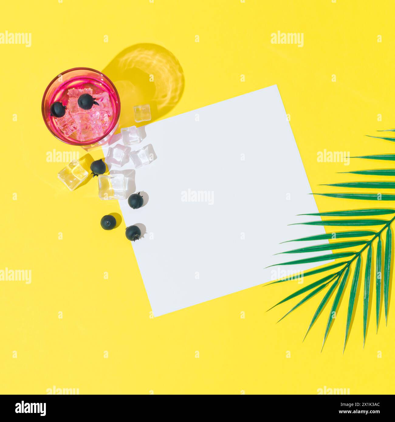 Summer layout made with cold refreshing drink, blueberries, ice cubes, tropical palm leaf and paper card note copy space on yellow background. Stock Photo