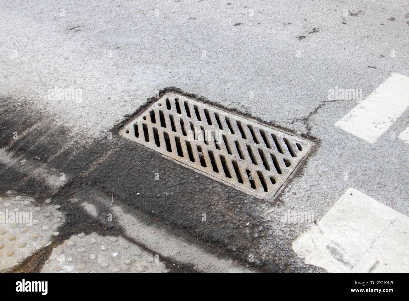A grey, rectangular manhole cover is resting on the asphalt road surface near a storm drain, possibly covering a sanitary sewer or gas line Stock Photo