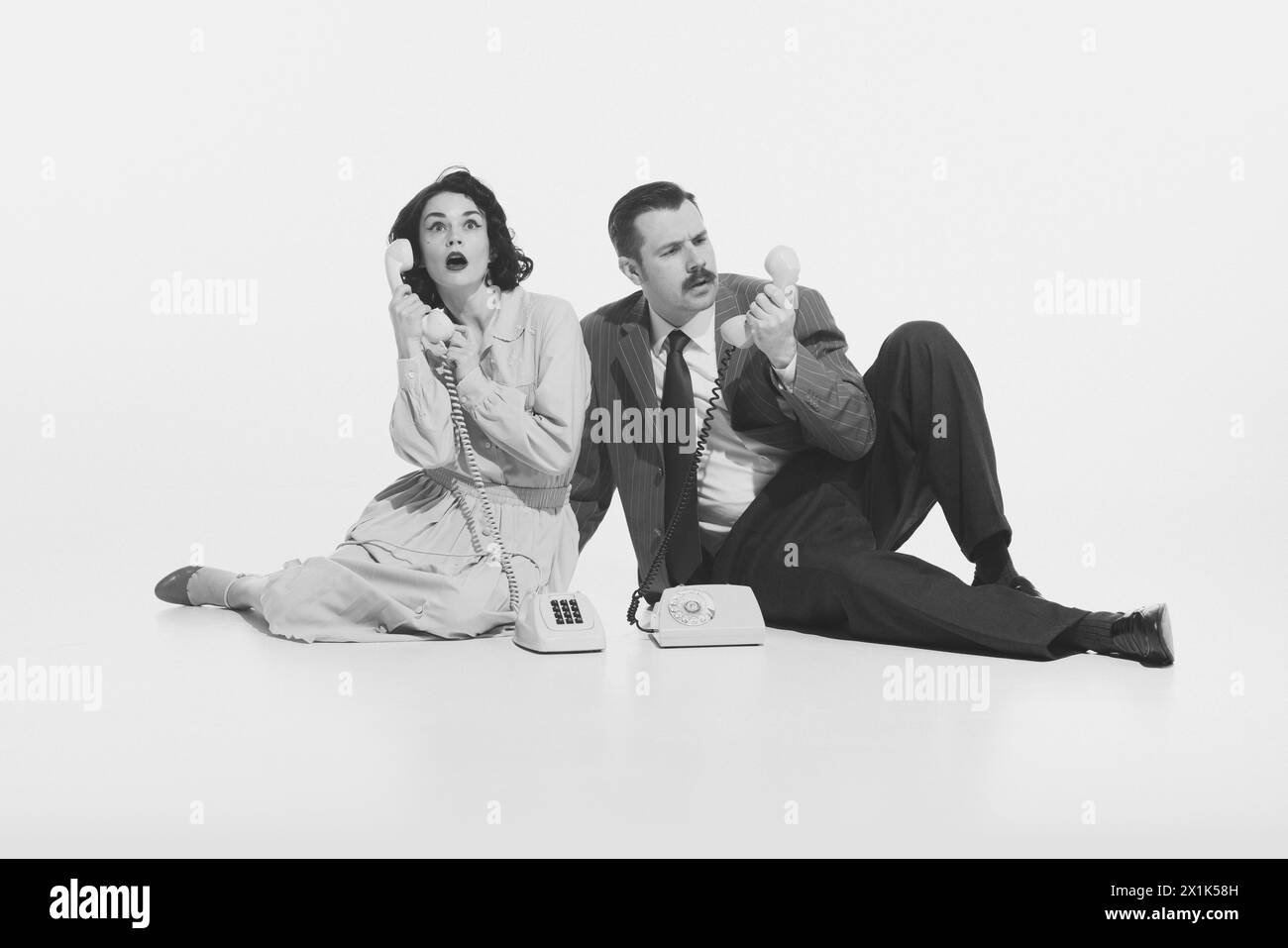 Monochrome i,age of elegant young woman and stylish man sitting on floor and expressively talking on vintage rotary phone Stock Photo