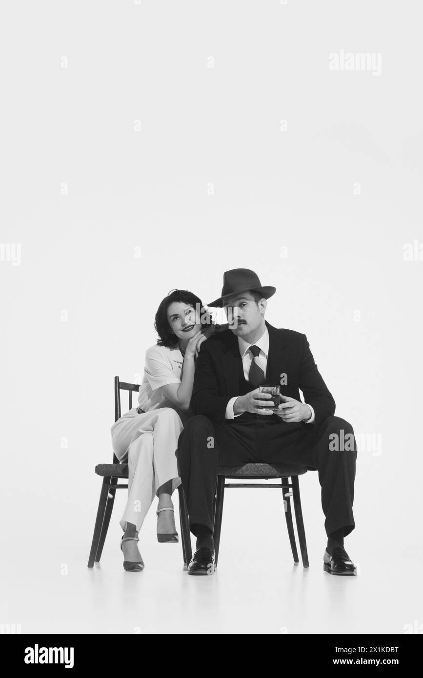 Monochrome portrait of young married couple, man in fedora hat and suit sitting with whisky glass and elegant smiling woman leaning on his shoulder Stock Photo