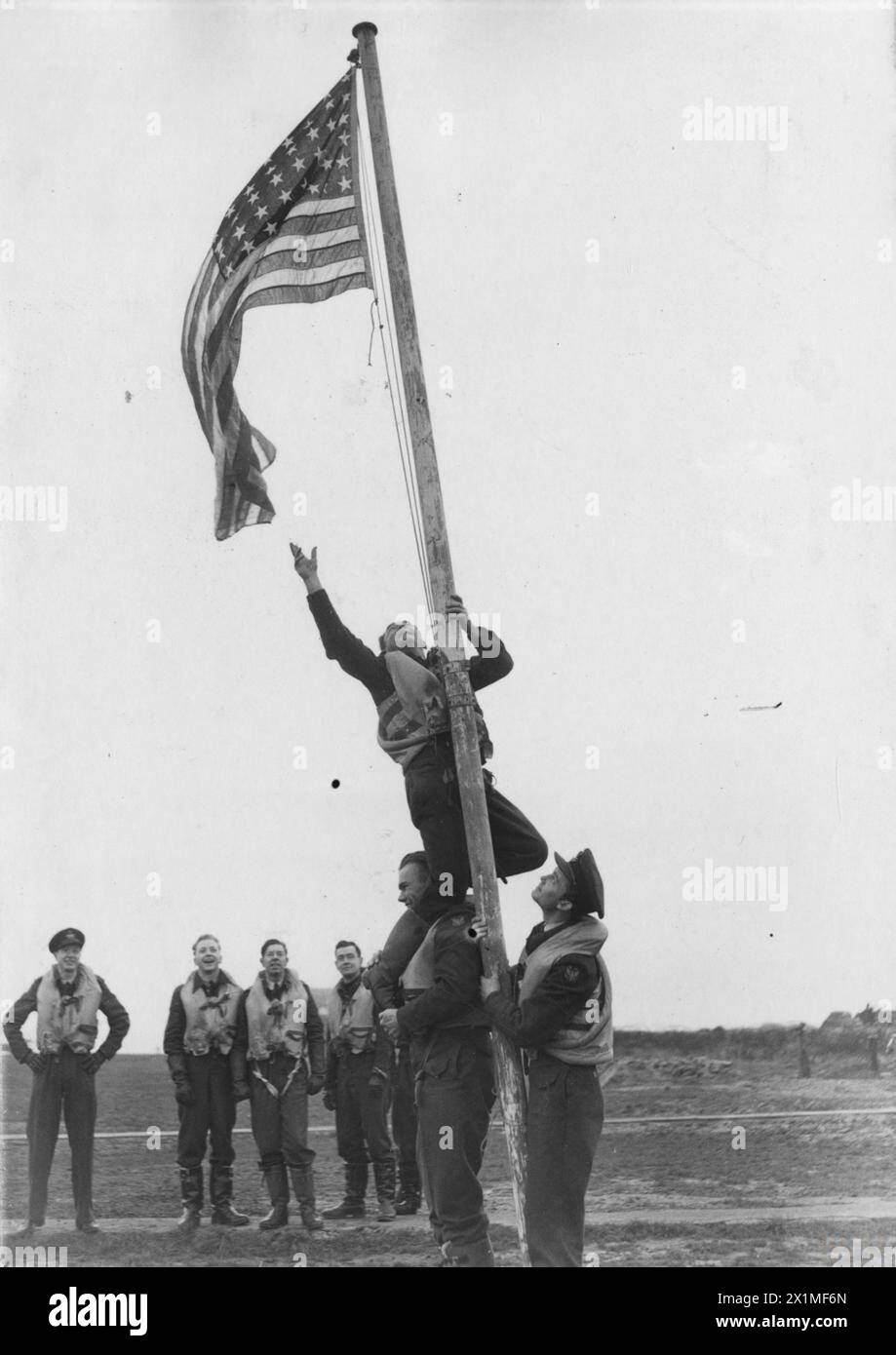 UNITED STATES EIGHTH AIR FORCE IN BRITAIN, 1942-1945 - Pilot Officer Kenneth Le Roy Holder stands on the shoulders of Pilot Officer Donald William McLeod to reach out for an American flag flying from a flag pole. A group of American airmen look on. They wear the insignia of the RAF Eagle Squadrons, the forerunners of the squadrons of the 4th Fighter Group. Passed for publication on 27 November 1941.Printed caption on reverse: 'Pilot Officer Kenneth Le Roy Holder from Buena Park, California, stands on the shoulders of Pilot Officer Donald William McLeod from Blackstone, Massachusetts, to fix th Stock Photo