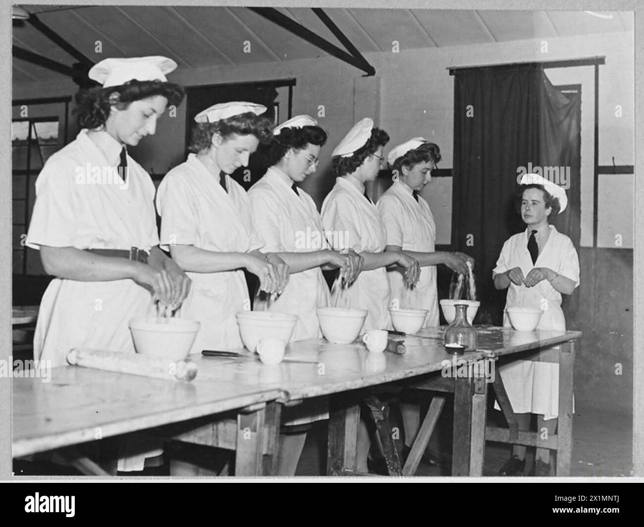 'CIVVY STREET COURSE' IN R.A.F. PATHFINDER GROUP - 16004 Picture issued 1945 shows - W.A.A.Fs learn to become good cooks during E.V.T. instruction. All R.A.F. and W.A.A.F. personnel are allowed 6 hours weekly of their working time for E.V.T. and any voluntary time they care to add. There is no rank distinction among pupils, all officers and airmen are referred to as 'Mr.'. W.A.A.F. officers and airwomen as 'Miss'. Little instructress [at end of line] with big job is Section Officer Berry Young of Jordan Hill, Glasgow. Catering Officer and expert in domestic science. She is showing the pupils, Stock Photo