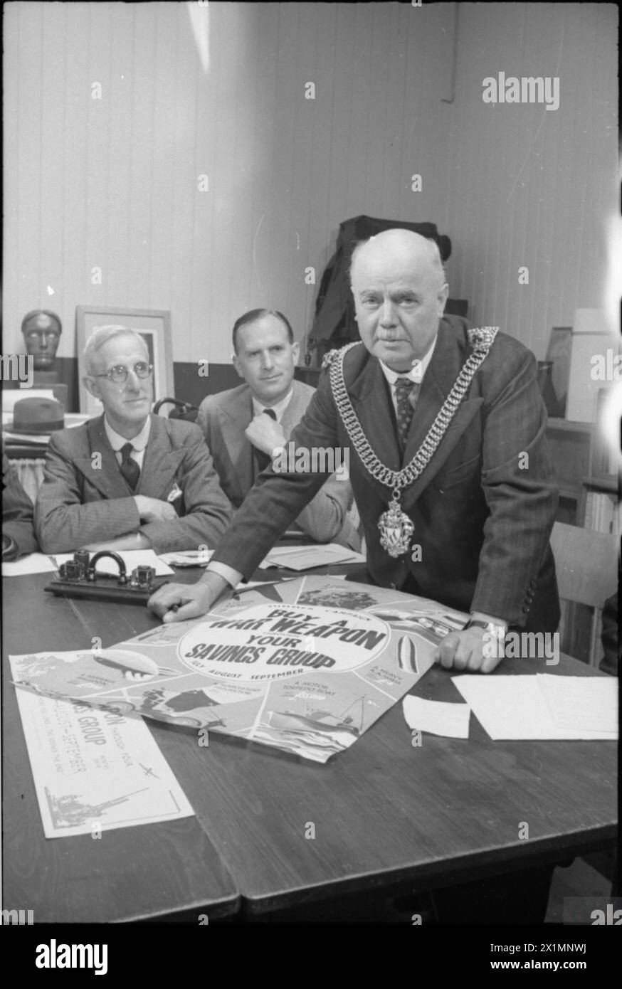 THE NATIONAL SAVINGS SCHEME AT WORK IN CANTERBURY, ENGLAND, 1941 - At the Sydney Cooper School of Art, St Peter's Street, Canterbury, a meeting was held to discuss plans for Canterbury's National Savings Campaign. At this meeting, Canterbury's 'Brain's Trust' was chaired by His Worship Alderman Charles Lefevre OBE, seen here leaning on the table. Also in attendance were Sydney Steele, Headmaster of Blean School (pictured on the left) and F E Clarke (centre), a local bank manager and Honorary Secretary of the Savings Committee. On the table, a large National Savings poster can be seen, Stock Photo