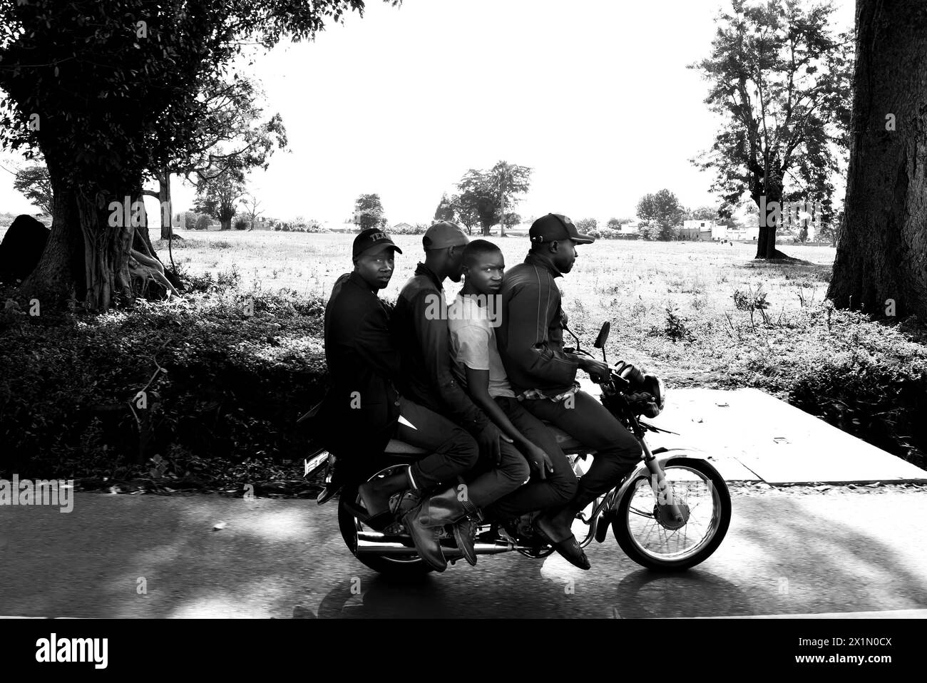 Youthful adventure: Four friends ride a motorcycle along a Ugandan road in the morning, one gazes at the camera. Black and white shot from a car, with Stock Photo