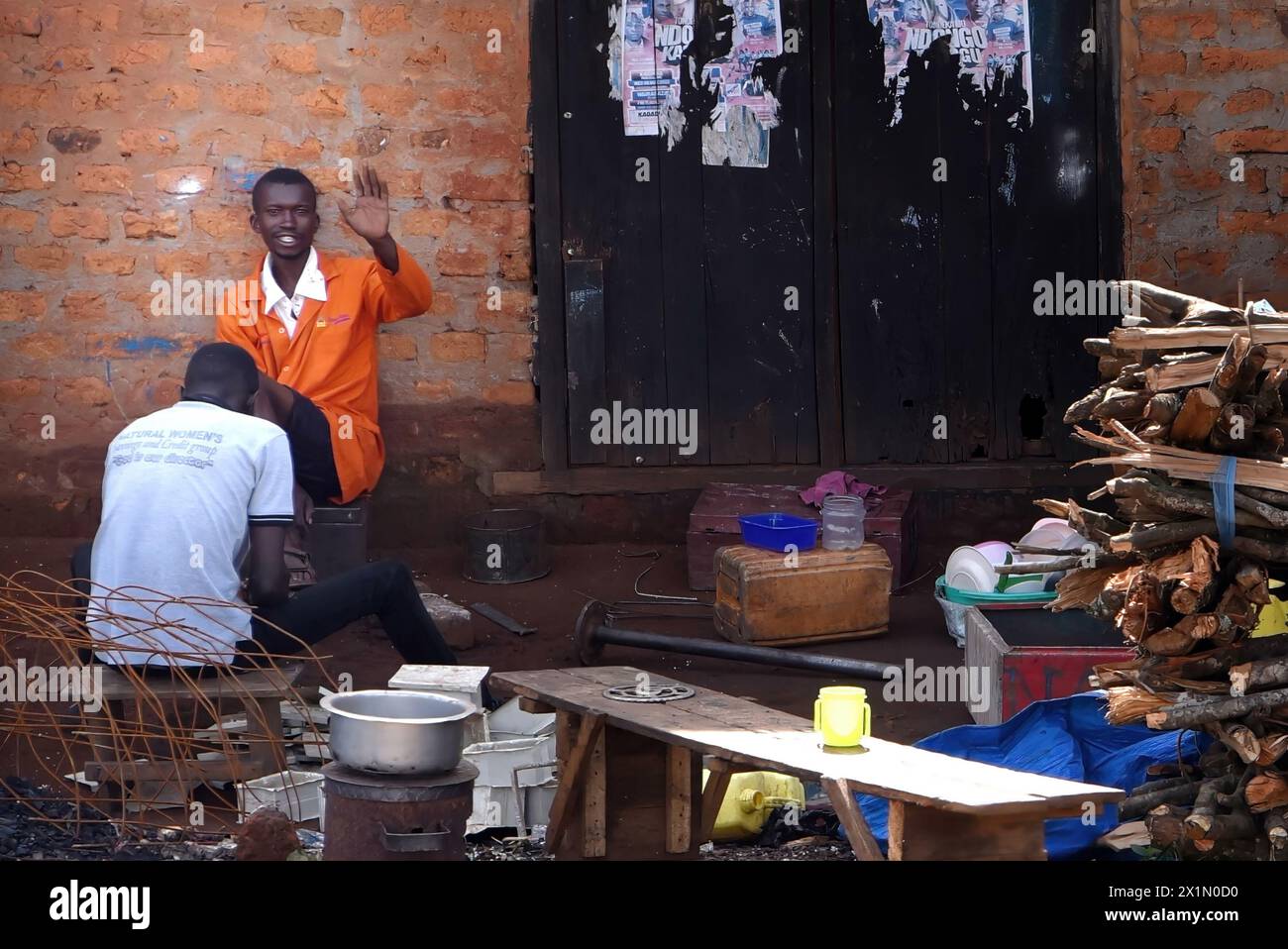 A warm greeting from Uganda's Masindi road: Amid scrap, a person smiles as another receives a pedicure, showcasing roadside hospitality and friendly c Stock Photo