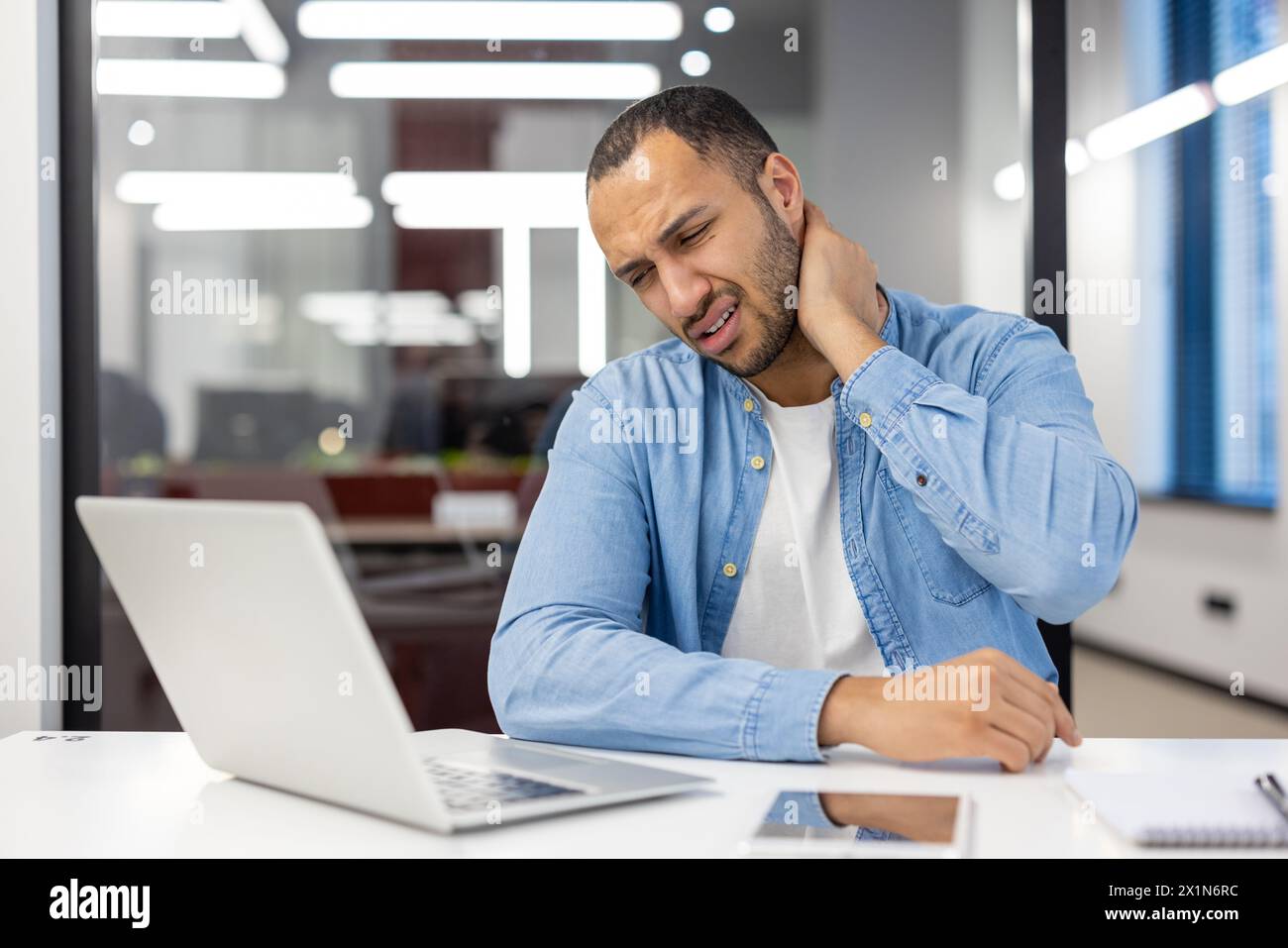 Tired hispanic man in shirt sitting in office at desk with laptop and holding hand on neck, doing massage, feeling pressure and pain. Stock Photo