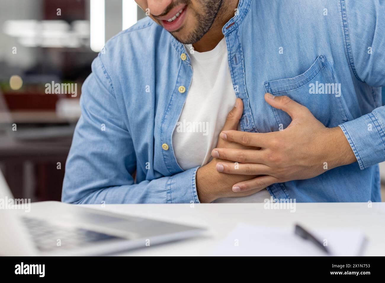 A close-up photo in the working office of a young man's hands holding his chest, feeling a strong pain in his heart. Stock Photo