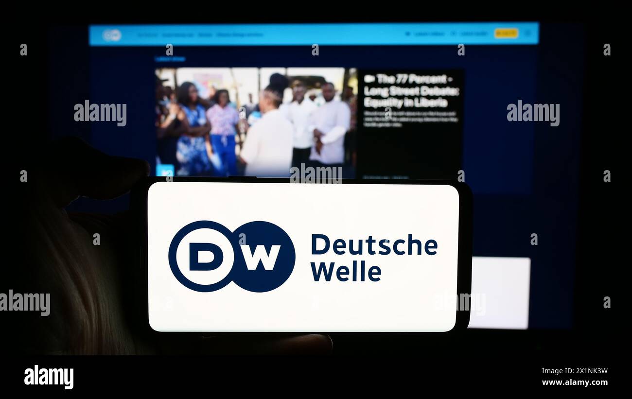 Person holding cellphone with logo of German state-owned broadcaster Deutsche Welle (DW) in front of webpage. Focus on phone display. Stock Photo