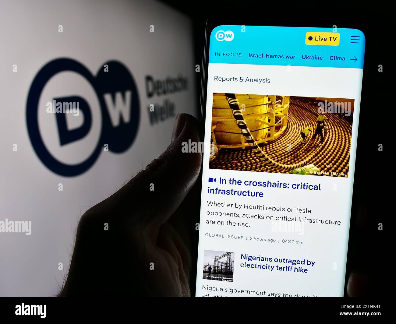 Person holding cellphone with webpage of German state-owned broadcaster Deutsche Welle (DW) in front of logo. Focus on center of phone display. Stock Photo