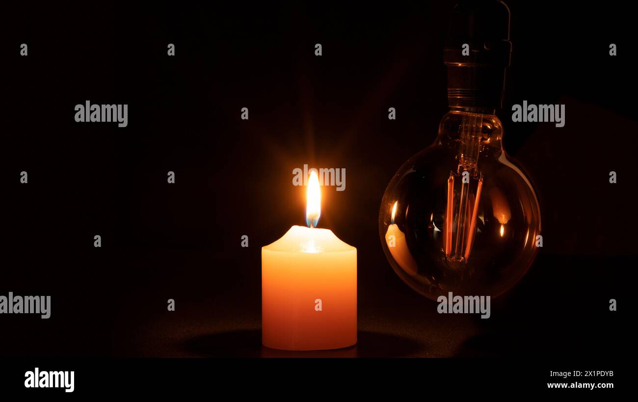 Blackout concept, turned off light bulb with no electricity hanging next to a burning candle against black background Stock Photo