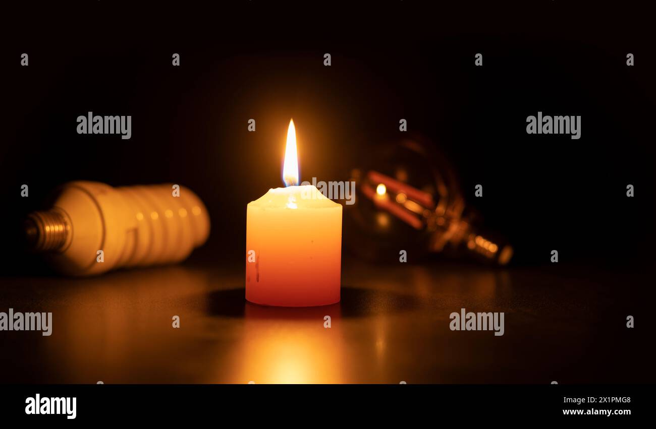 Blackout concept, A white burning candle in close-up on a black surface against two bulb lights in black background Stock Photo