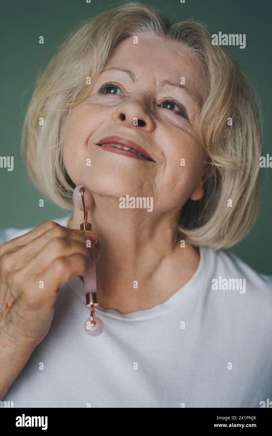 Elderly woman massaging her face and neck with roller made with natural stone. Keeping youth, take care of skin, pamper yourself. Skincare, wellness Stock Photo