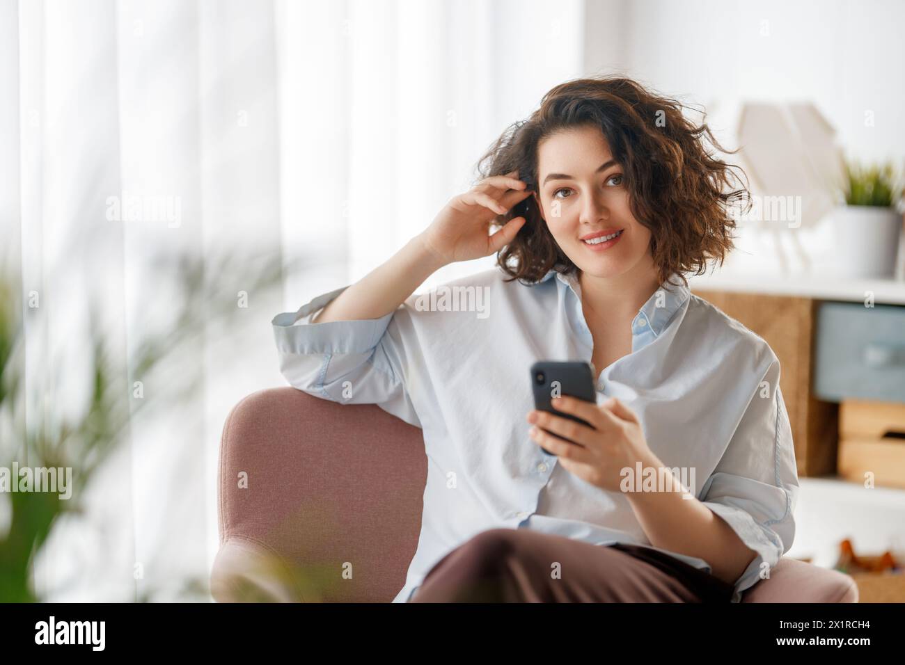 Happy casual beautiful woman is using a phone sitting on armchair at home. Stock Photo