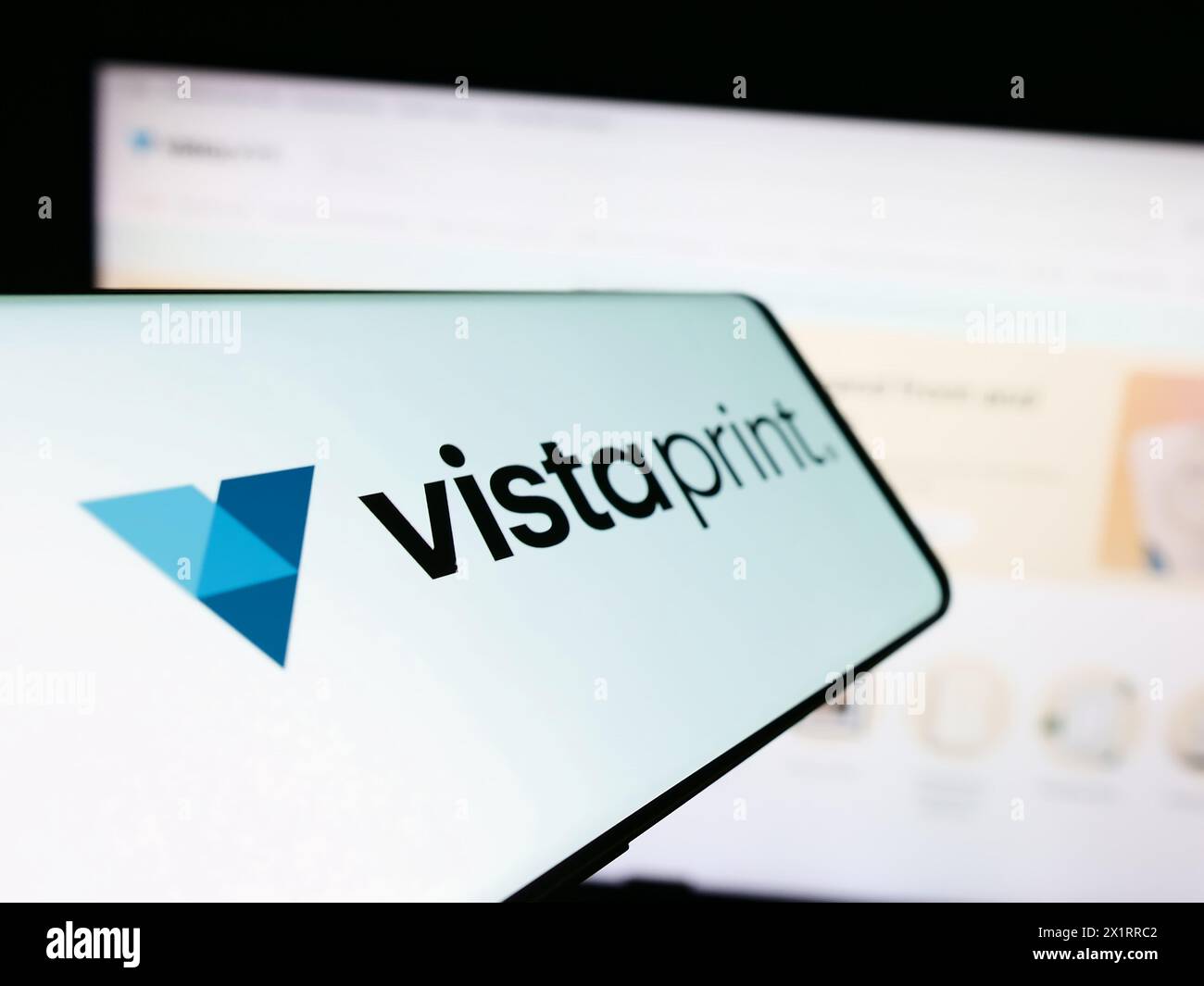 Mobile phone with logo of mass customization company VistaPrint (Vista) in front of business website. Focus on center-left of phone display. Stock Photo