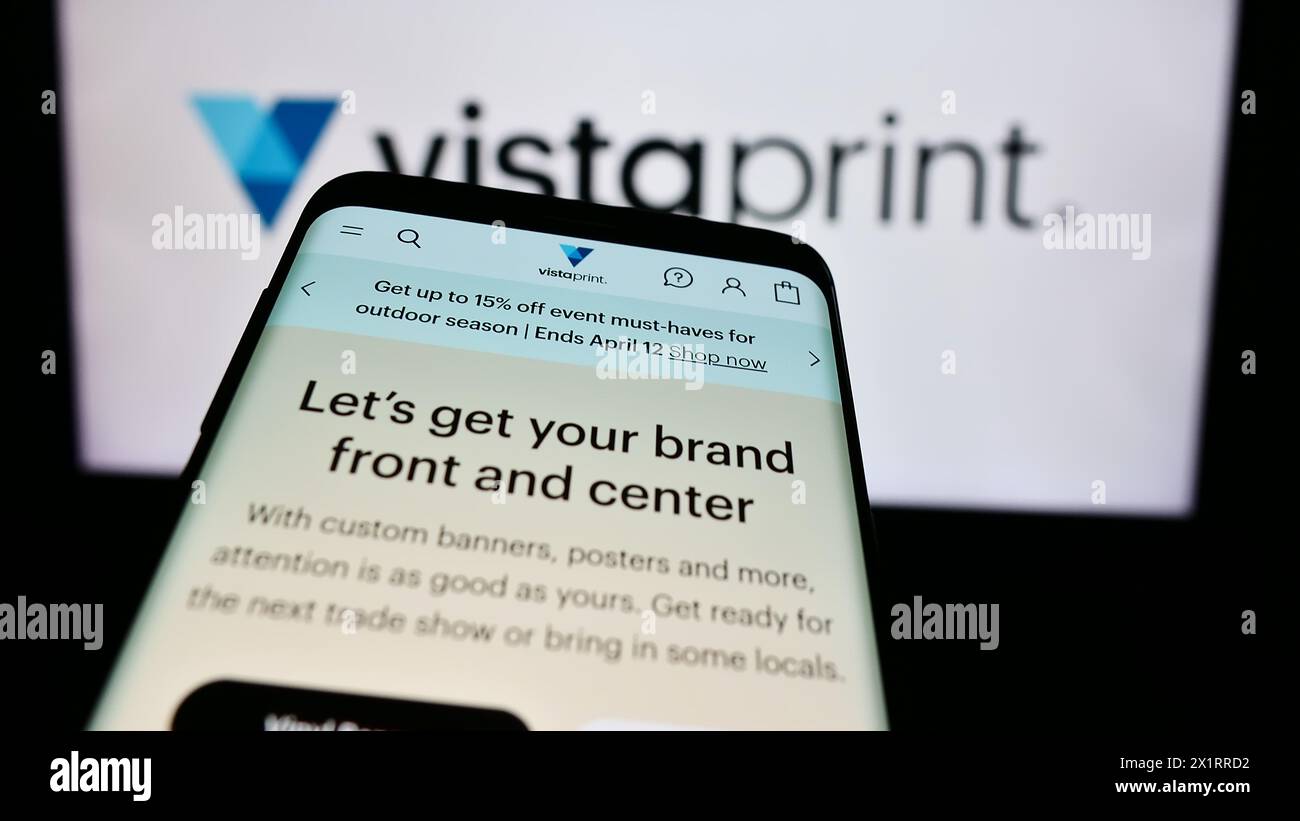 Smartphone with website of mass customization company VistaPrint (Vista) in front of business logo. Focus on top-left of phone display. Stock Photo