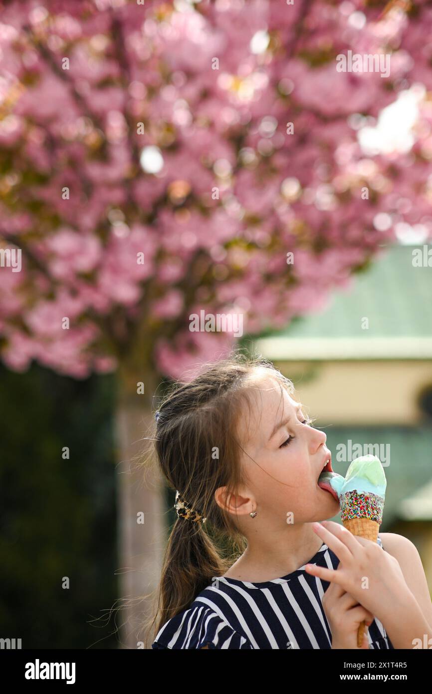 Little girl licks ice cream in summer weather. Beautiful pink blooming tree in the background-bokeh. Stock Photo