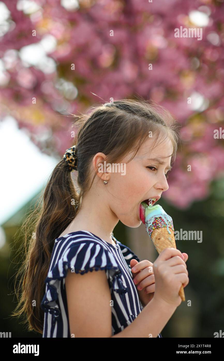 Little girl licks ice cream in summer weather. Beautiful pink blooming tree in the background-bokeh. Stock Photo