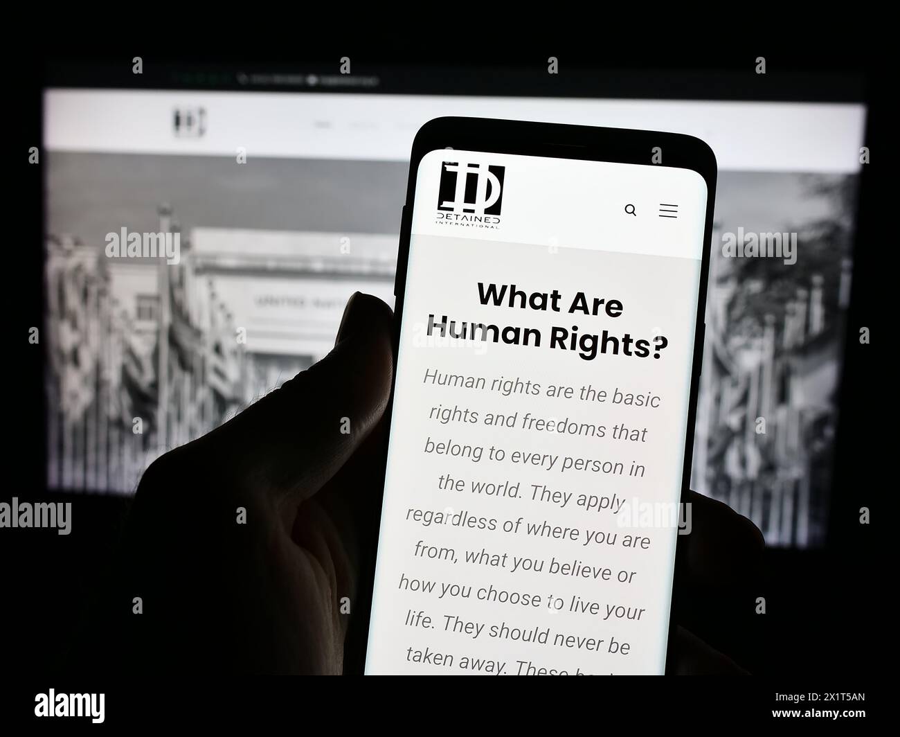 Person holding cellphone with webpage and logo of legal advocacy NGO Detained International in front of monitor. Focus on center of phone display. Stock Photo