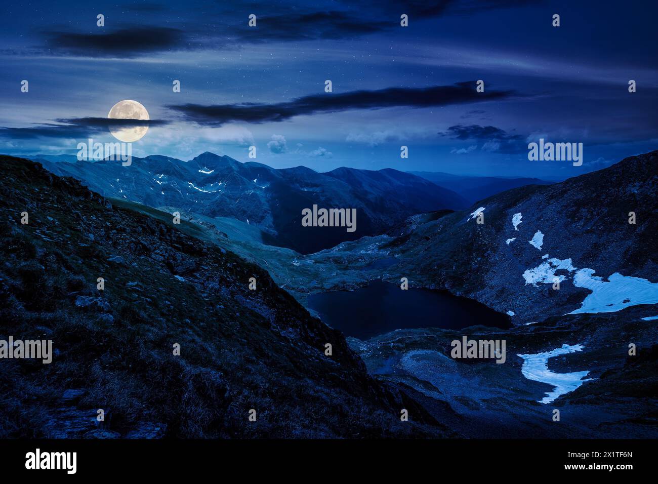 alpine landscape of fagaras mountains at night. capra lake of romania in summer. spots of snow and grass on the rocky hillside in full moon light. stu Stock Photo