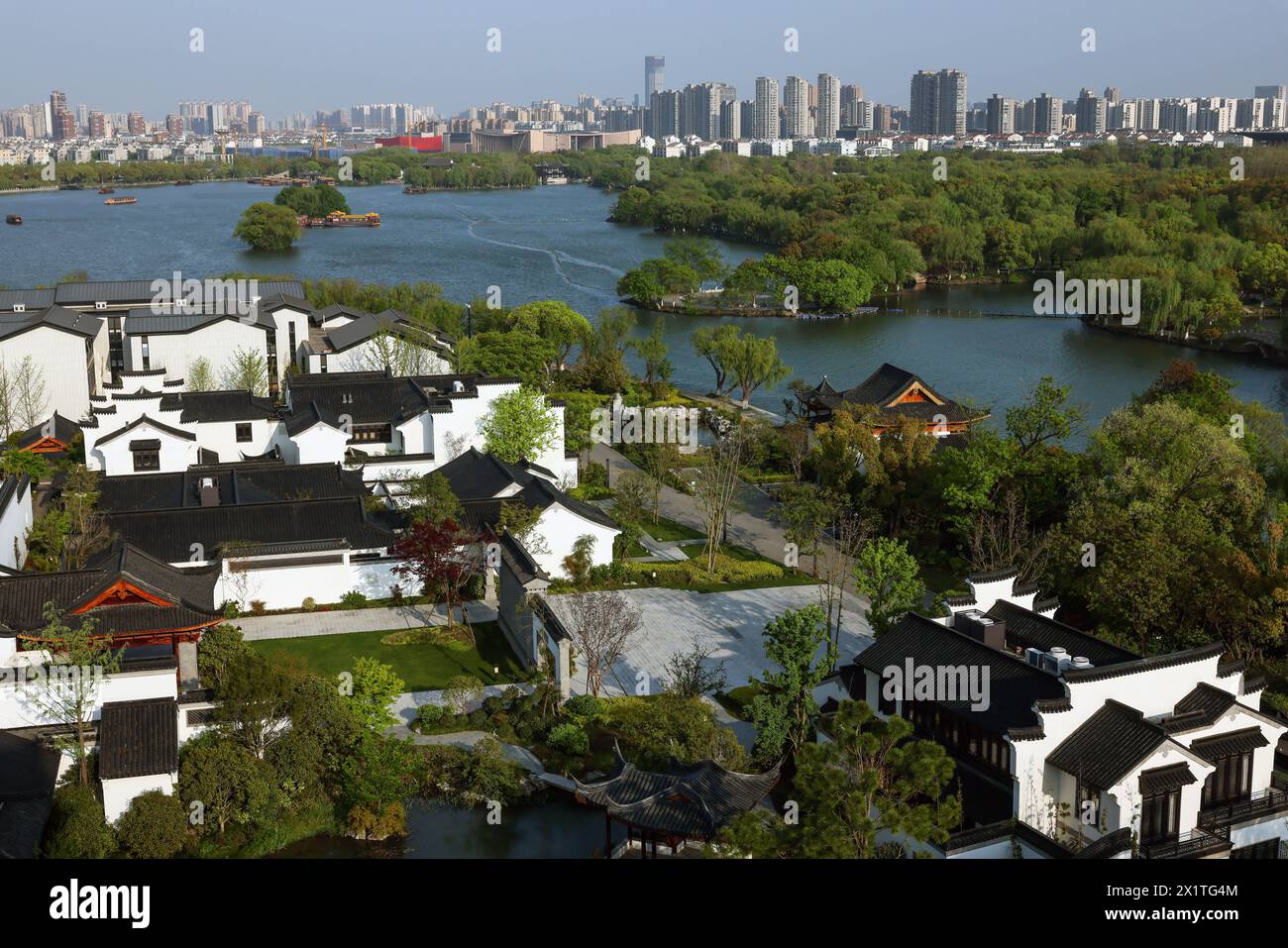 (240418) -- JIAXING(ZHEJIANG), April 18, 2024 (Xinhua) -- Ancient dwellings are seen against the distant city skyline near the Nanhu Lake in Jiaxing City, east China's Zhejiang Province, April 10, 2024. Located in east China's Zhejiang Province, the waterside city Jiaxing is renowned for tourist destinations Nanhu Lake and Wuzhen. In the breezy month of April, the two tourist hotspots come alive with the gentle touch of spring. Nanhu Lake, with its shimmering waters and willow trees swaying along the lakeside, is a sight to behold. The historic water town Wuzhen also enchants with its emer Stock Photo