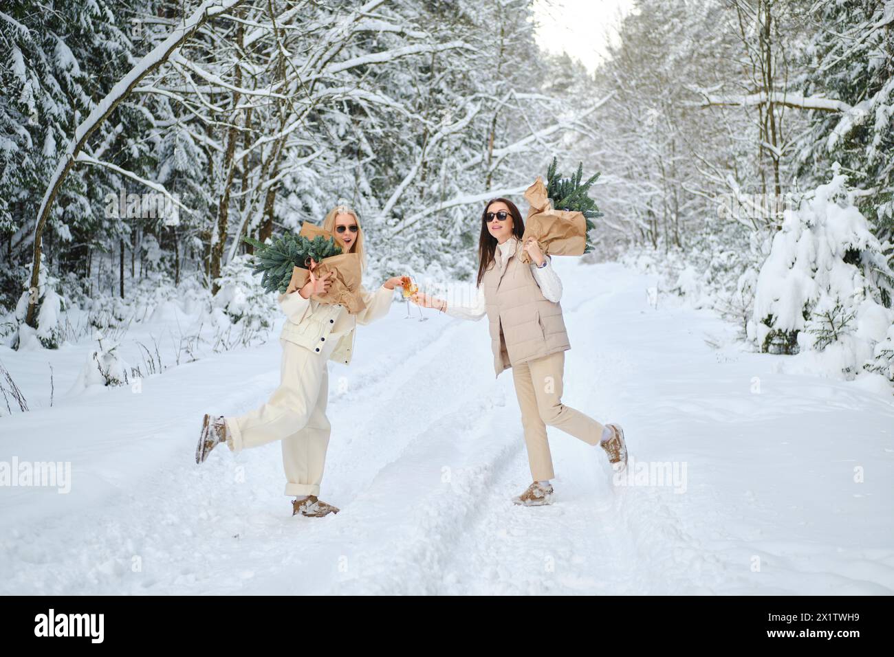 Two joyful women in snowy forest holding fir tree branches on their shoulders and clinking glasses with champagne Stock Photo