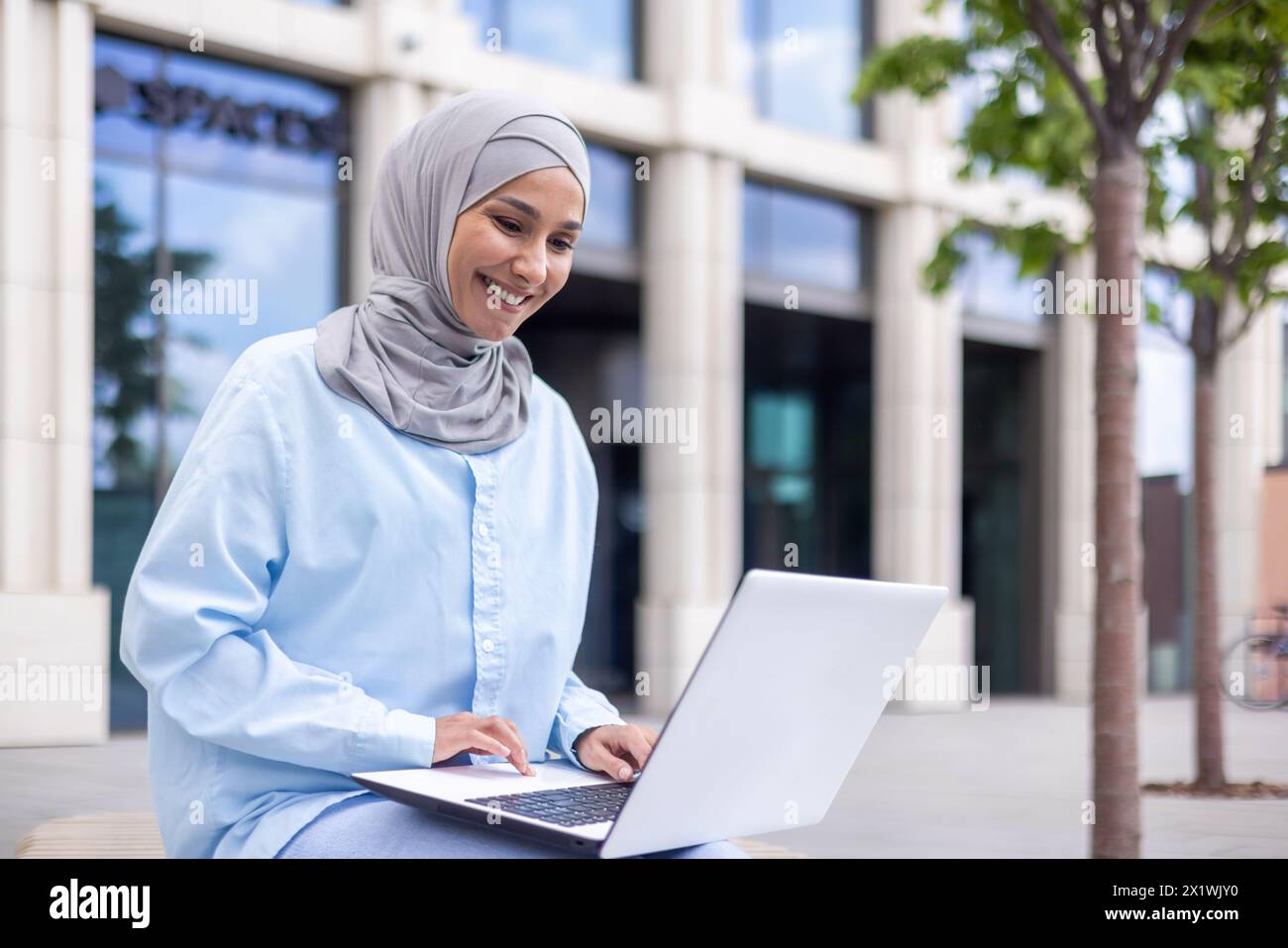A cheerful Muslim woman in a blue hijab works on her laptop while sitting outside a contemporary building. She is enthusiastic and focused, showcasing diversity and modernity in work environments. Stock Photo