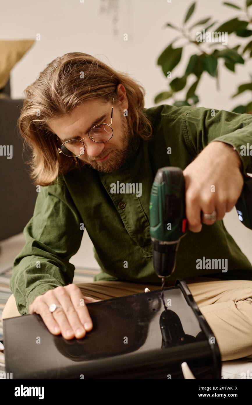 Young man in eyeglasses sitting on the floor and bending over plastic bucket while making several holes with electric handtool Stock Photo