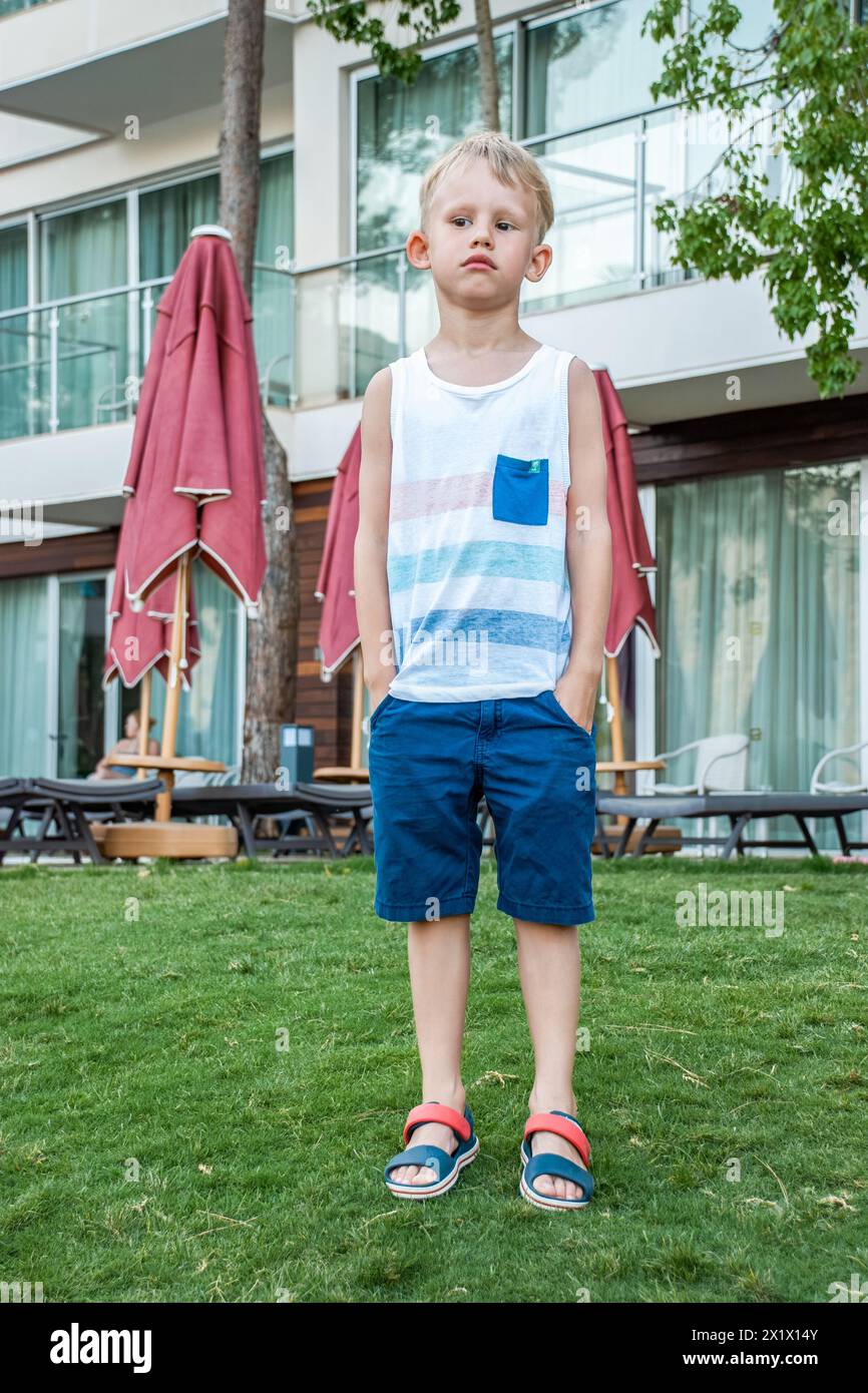 Sad boy in beach clothes stands on a well-maintained lawn on background of modern seaside hotel with closed red umbrellas and lounge chairs Stock Photo