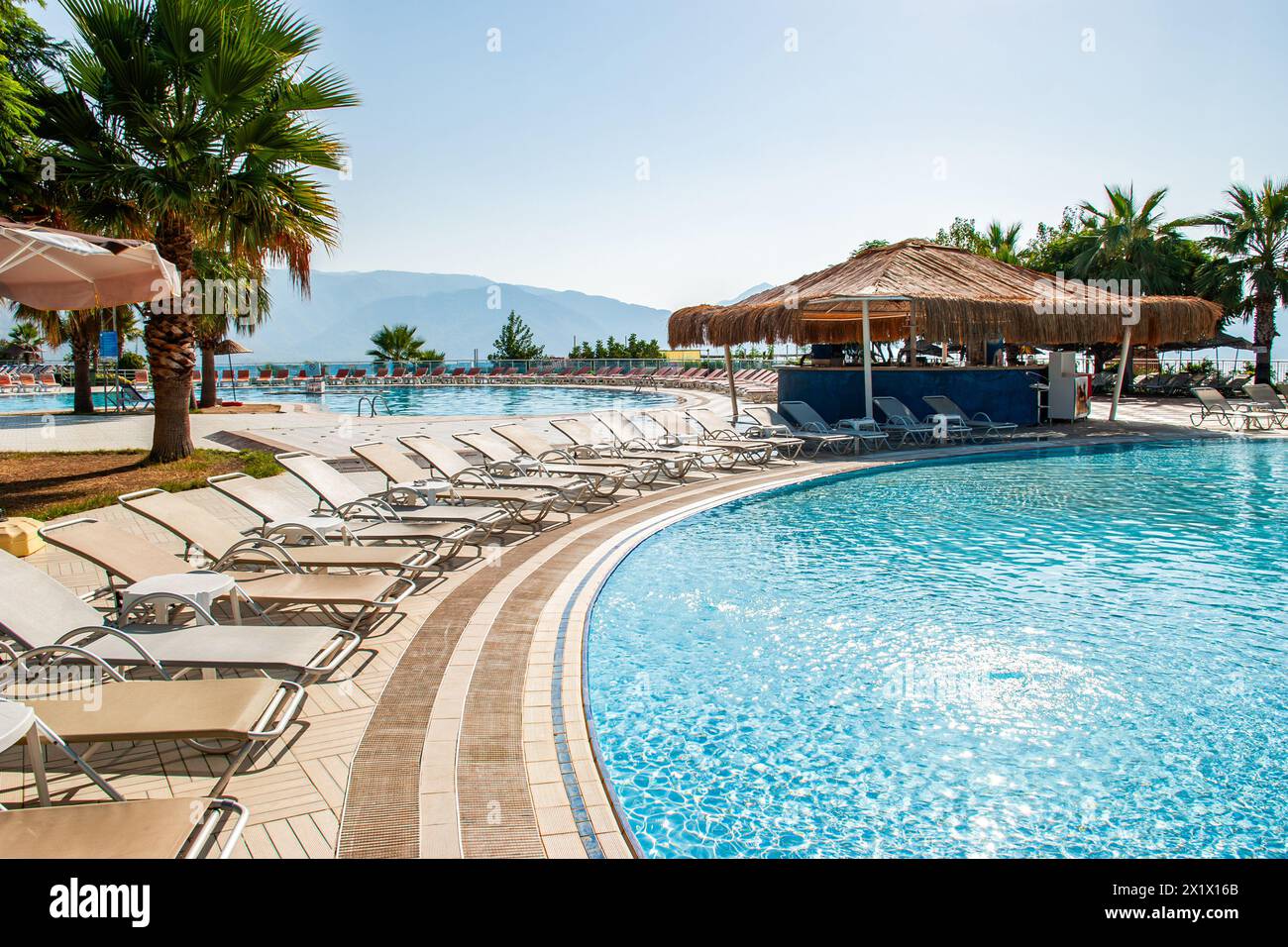 Circular swimming pool surrounded by lounge chairs and a bar with a thatched roof, in a summer water park in the early morning Stock Photo