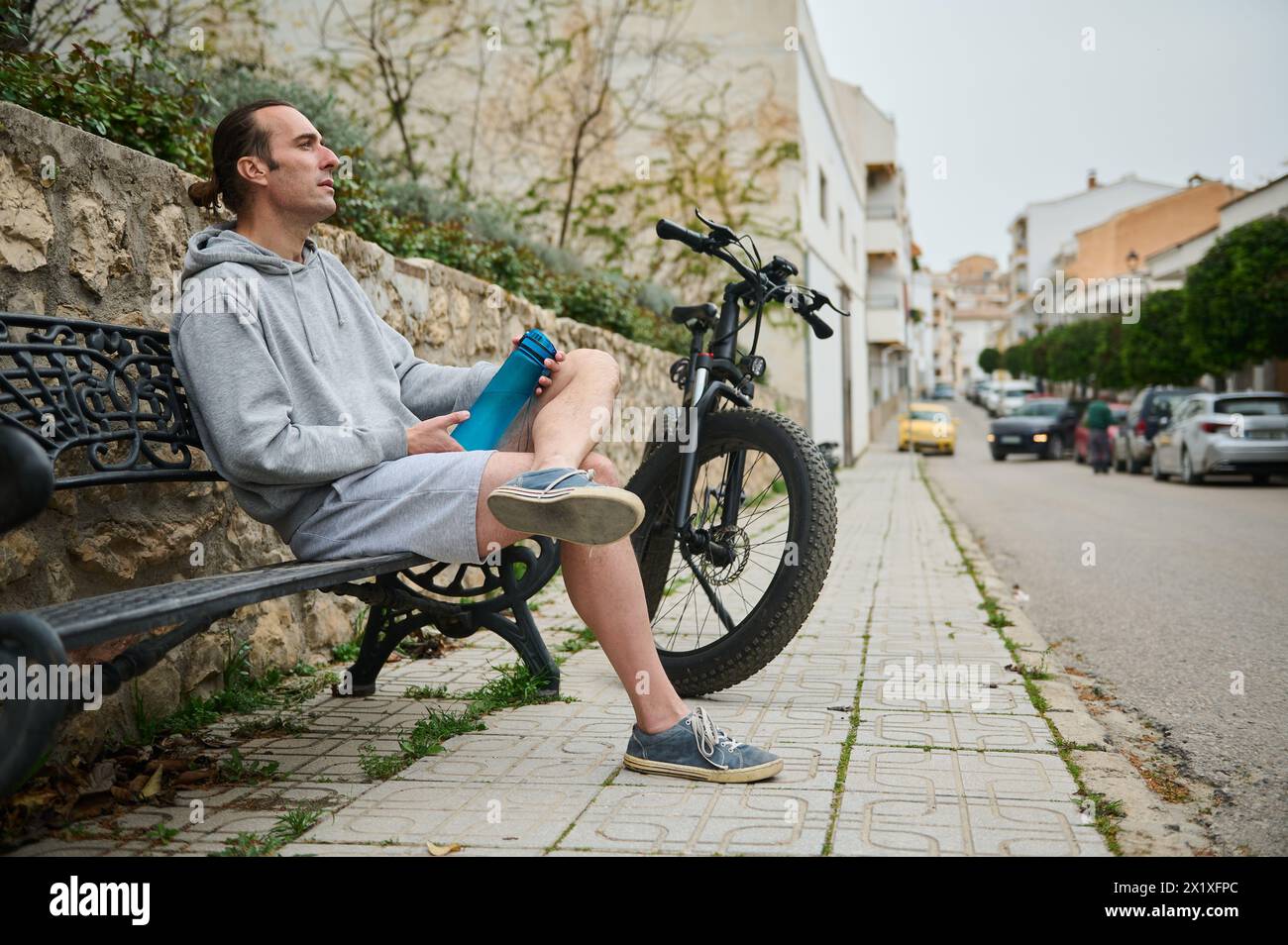 Caucasian young hipster sporty man holding a a sports bottle of pure water, sitting on a bench near his electric battery powered motor bike Stock Photo