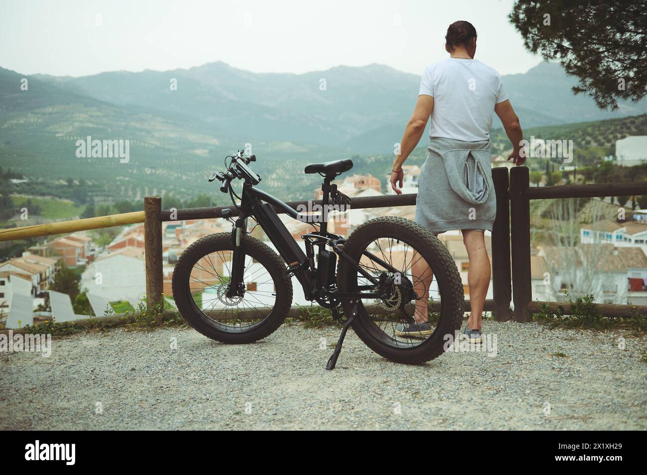 Rear view of a young man cyclist, standing near his electric bike, having a break after biking in mountains. People. Active healthy lifestyle. E-bike Stock Photo