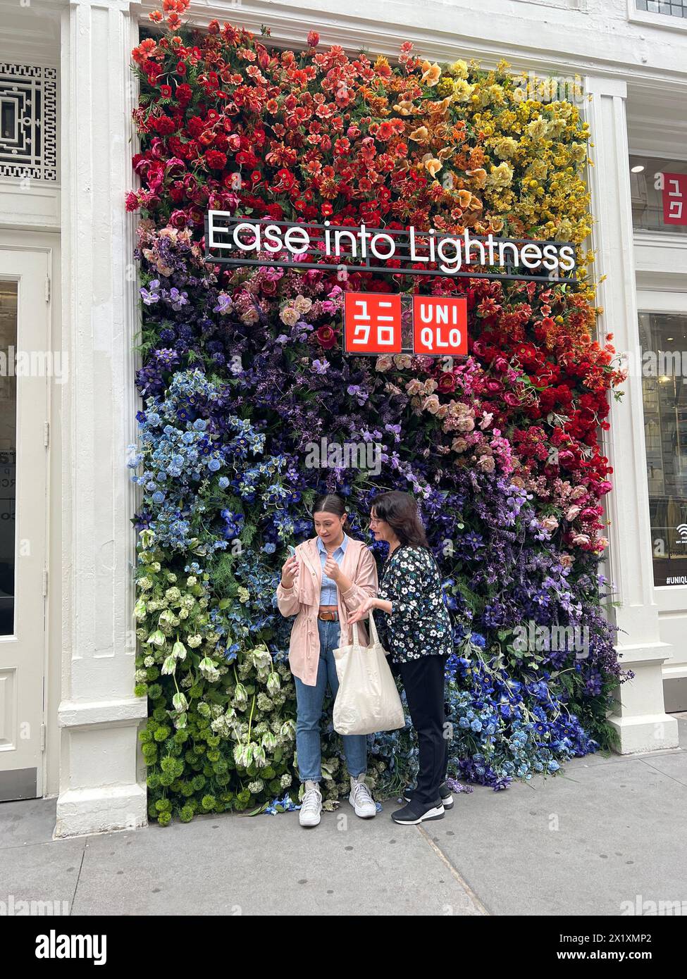 Women looking at their phone screen in front of a flowered wall at the Uni Clo store on Broadway in the SOHO, neighborhood of Manhattan, NYC. Stock Photo