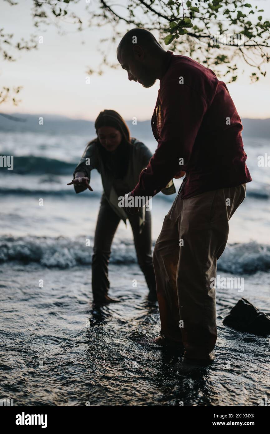 Silhouetted man helping a woman steady herself in rough lake waves at sunset Stock Photo