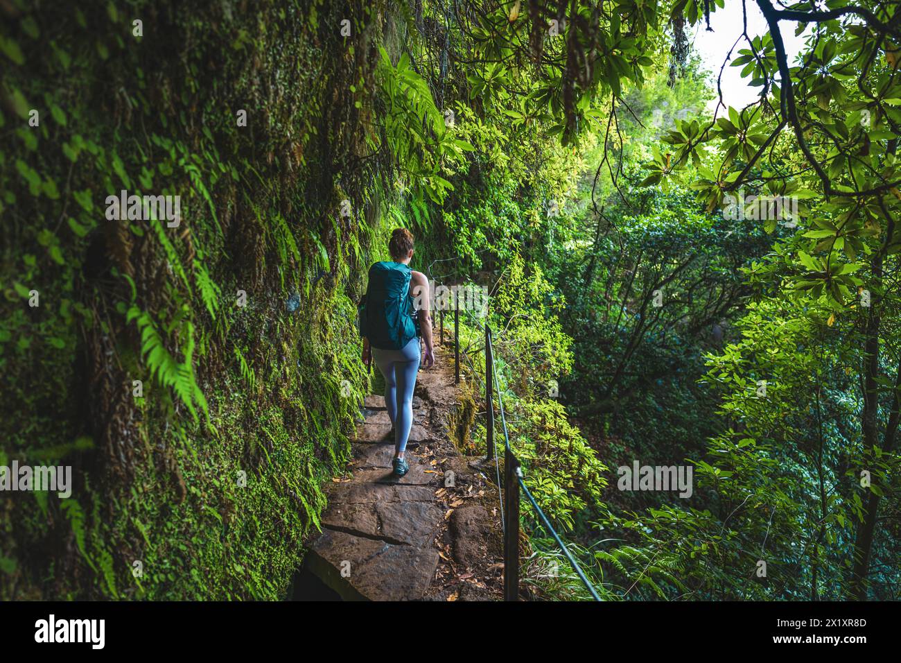 Description: Female backpacker walking along rainforest water channel path on steep cliff covered with plants. Levada of Caldeirão Verde, Madeira Isla Stock Photo