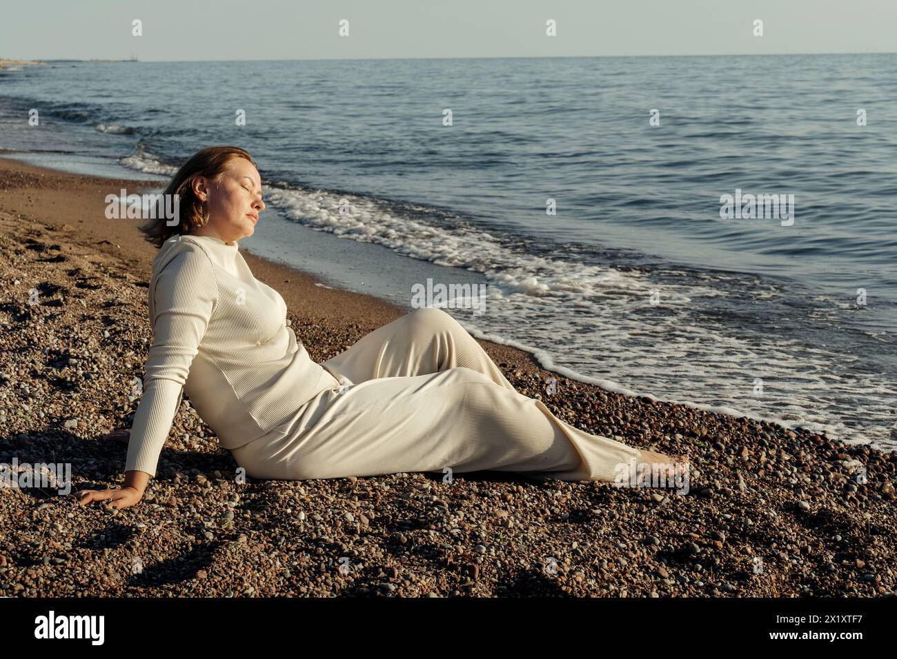 A woman is seated on a sandy beach, next to the ocean, with waves gently lapping the shore in the background. Stock Photo