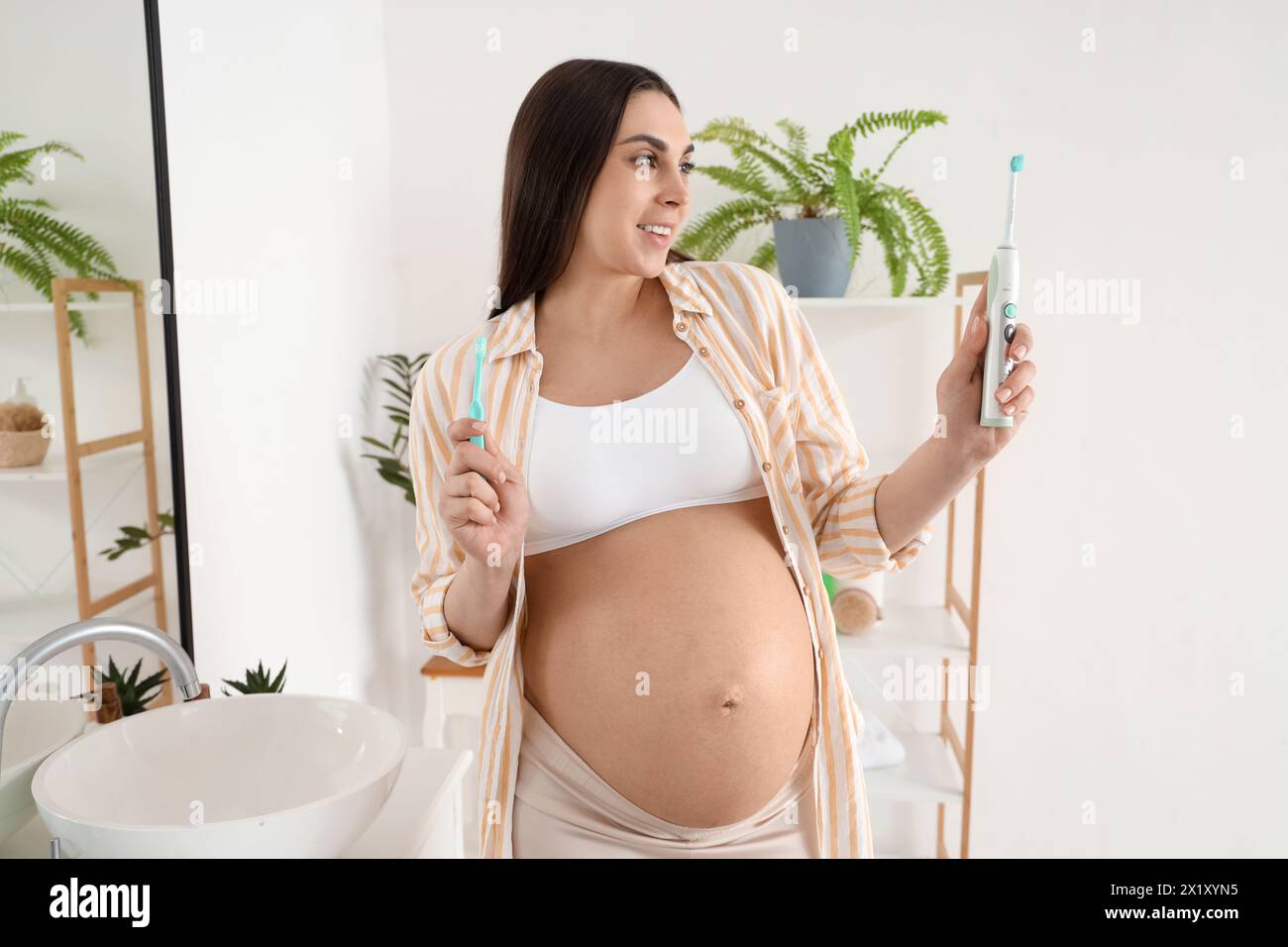 Young pregnant woman holding plastic and electric tooth brushes in white bathroom Stock Photo