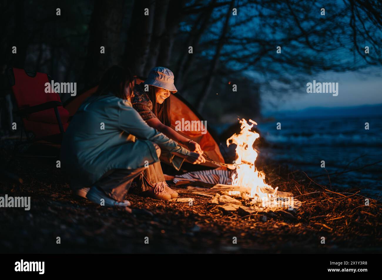 Friends enjoying a warm bonfire by the lakeside during a camping trip Stock Photo