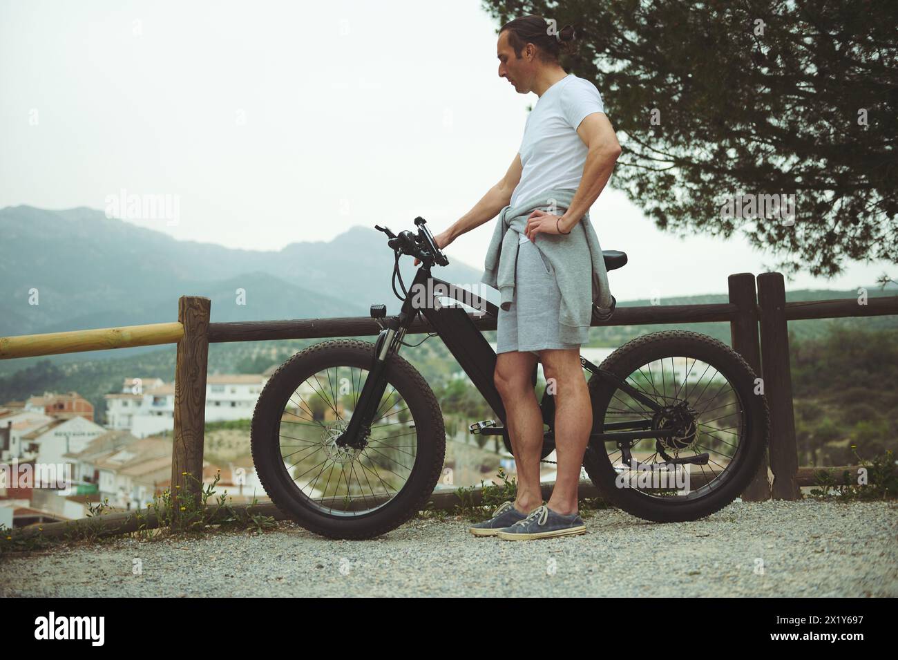 Full length European male cyclist relaxing on top of mountain, keeping hands on handlebar of his electric pedal-assist bicycle, enjoying amazing urban Stock Photo