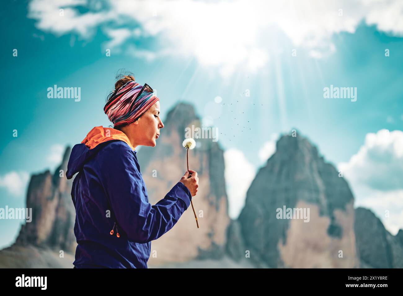 Description: Young woman blows seed from a dandelion in the afternoon. Tre Cime, Dolomites, South Tirol, Italy, Europe. Stock Photo