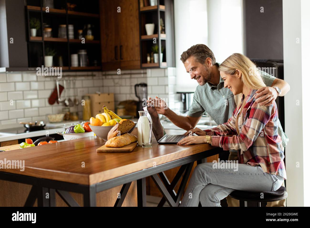 A cheerful couple enjoys a light-hearted moment in their sunny kitchen, working on laptop surrounded by a healthy breakfast Stock Photo