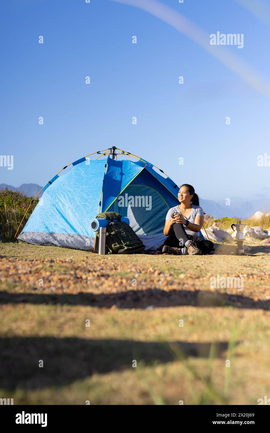 Biracial female hiker sitting near blue tent, enjoying nature, copy space Stock Photo