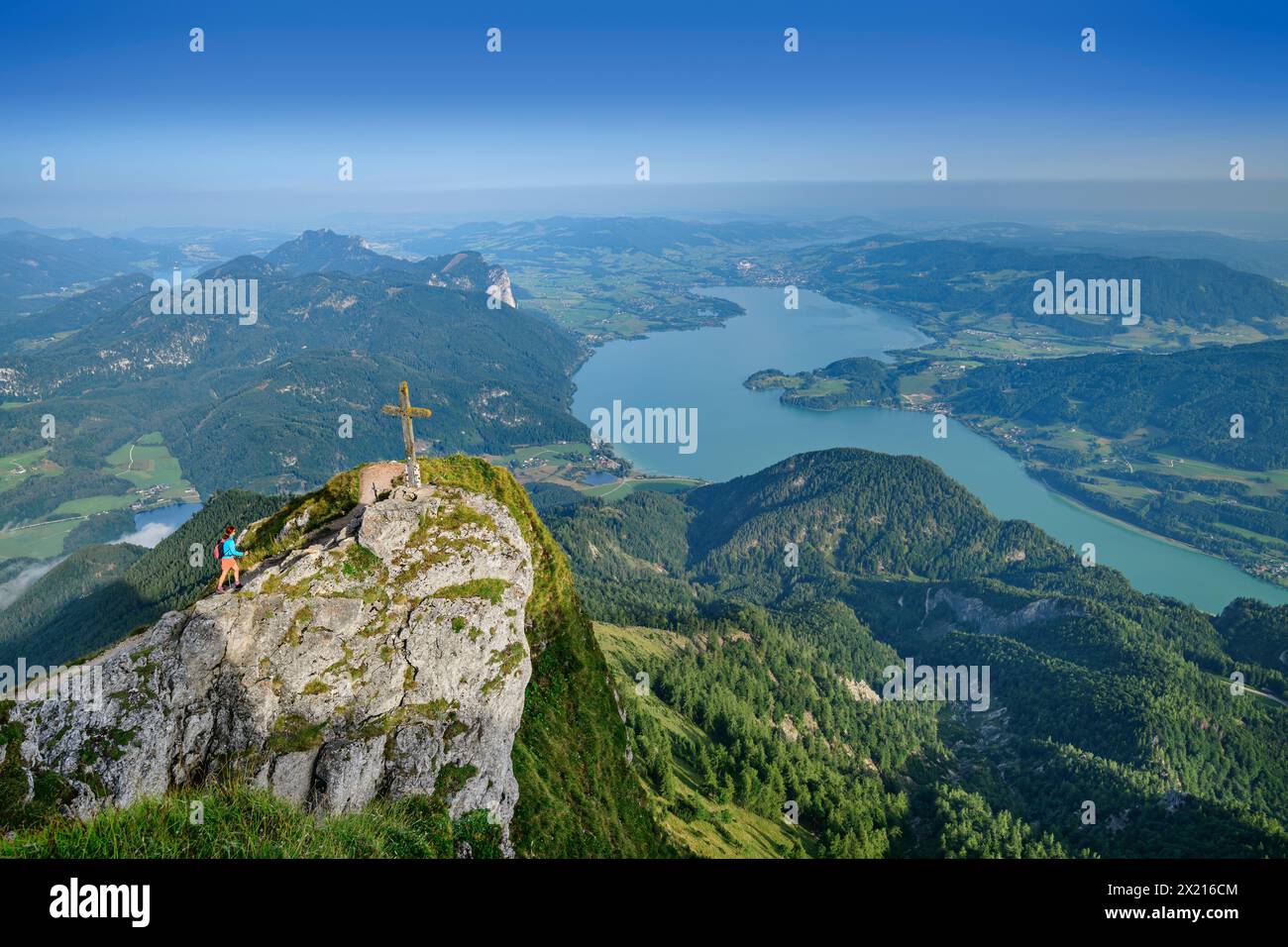 Woman hiking approaches the summit cross at the Himmelspforte, Mondsee in the background, Himmelspforte, Schafberg, Salzkammergut Mountains, Salzkamme Stock Photo