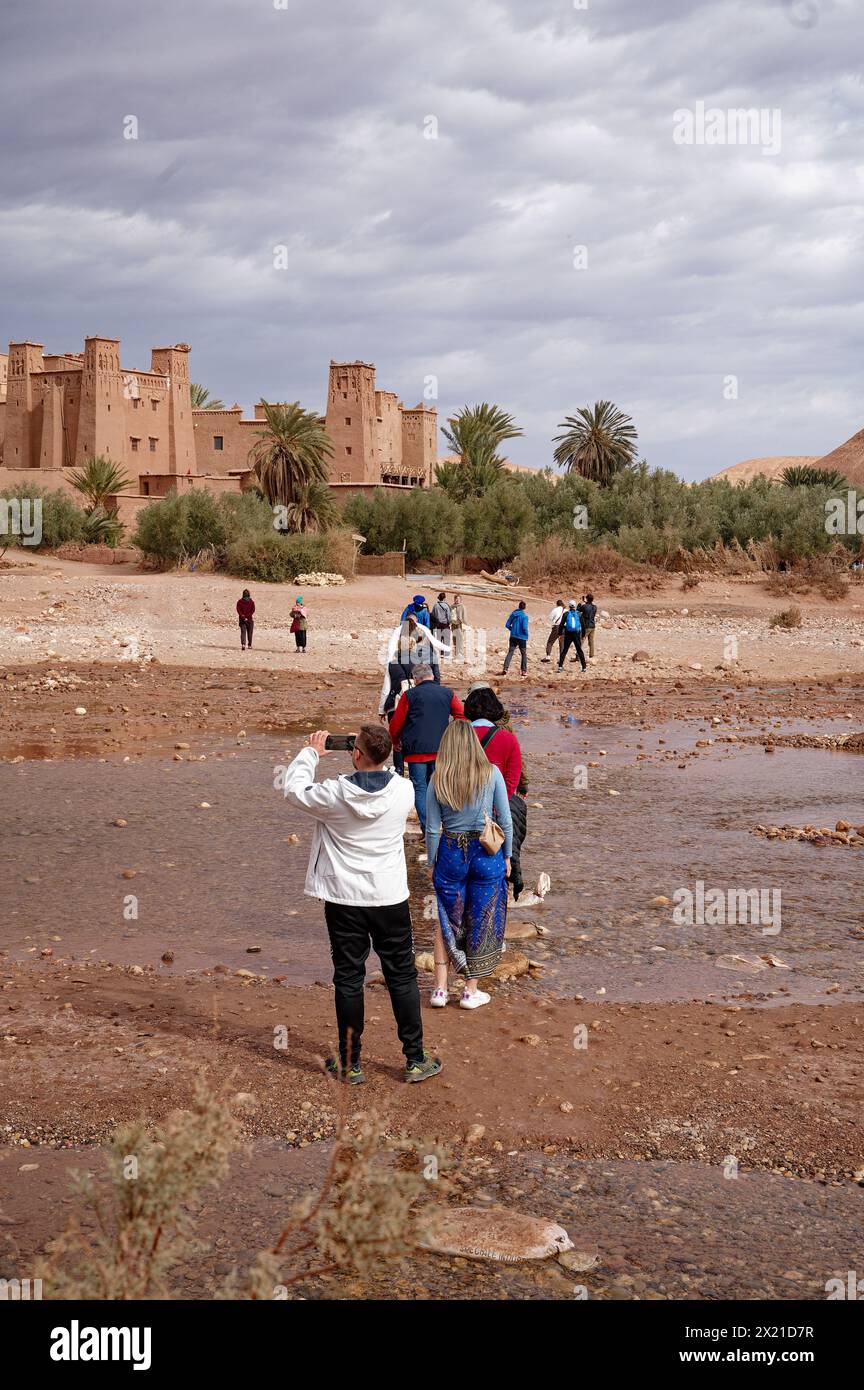 Tourists traverse a shallow river to the looming fortified village of Aït Benhaddou under a brooding sky. Stock Photo