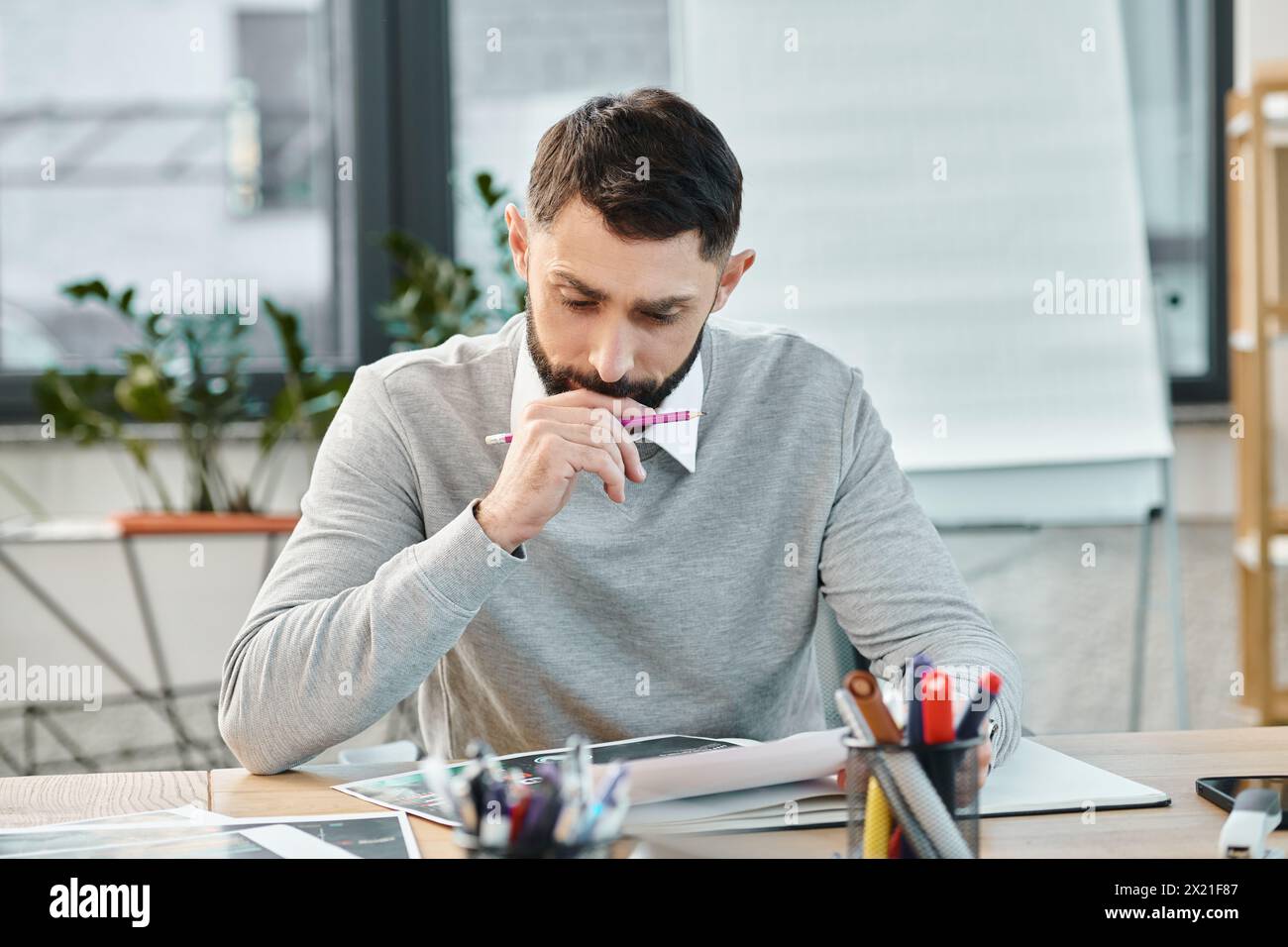 A man sits at a desk, engrossed in a piece of paper, deep in thought, surrounded by the hustle of a corporate office. Stock Photo