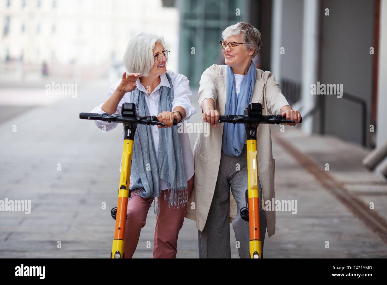 Elderly joyful gray haired women friends ride electric scooters in the city, happy smiling, enjoying a fun time together. Stock Photo